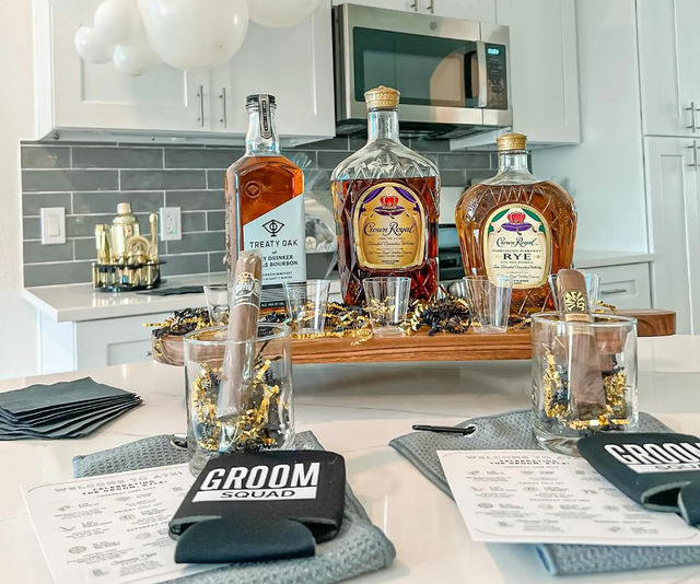 Groom-themed home kitchen drink station with three whiskey bottles on a wooden tray, shot glasses, two cigars in rocks glasses with gold confetti, 'Groom Squad' koozies and printed menus on a white island, white cabinets and gray subway tile backsplash with balloons overhead.