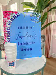 Blue welcome sign for a bachelorette weekend in Austin, TX with disco ball graphic and pink floral border, stacked white party cups on a marble table in a bright indoor setting