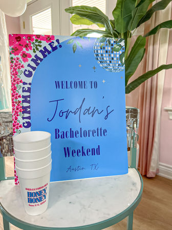 Bright blue welcome sign with disco-ball graphic and pink floral trim announcing a bachelorette weekend in Austin, TX, next to stacked white party cups on a marble-top table