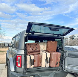 Gray SUV with rear hatch open in a suburban parking lot under a bright blue sky, trunk neatly packed with stacked beige and brown suitcases and travel bags, apartment buildings in the background.