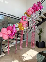 Playful rodeo-themed party decor: pink and gold balloon garland with cow-print balloons, pink cowboy-boot balloons, silver letter balloons spelling RODEO, and pink fringe streamers draped along a modern indoor staircase.