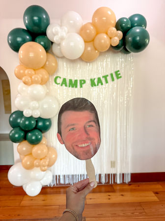Indoor camp-themed party backdrop with green, peach, and white balloon arch, green letter banner reading “CAMP KATIE,” shimmering fringe curtain, and a hand holding a smiling face photo-on-stick prop.