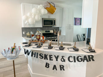 Modern white kitchen island converted into a whiskey & cigar bar for a celebration — whiskey bottles and glasses, cigar place settings labeled "GROOM", ice bowl with beers and champagne, and a beer-mug balloon cluster.