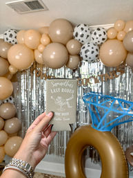 Hand holding a beige drink koozie labeled Last Rodeo with June 2025 and Austin, Texas, against a neutral tan balloon garland, silver fringe backdrop, large inflatable ring and blue diamond prop — playful bachelorette party decor.