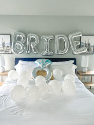 Silver foil 'BRIDE' balloons strung above a modern bedroom bed with an inflatable engagement-ring balloon and scattered white balloons on crisp white sheets, cactus wall art and bedside lamps — bachelorette party decor.