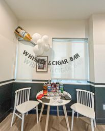 Whiskey-and-cigar bar party corner with banner and giant beer balloon above a small round table holding bottles, playing cards and two white chairs