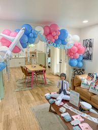 Airplane-themed baby shower in a bright living room with pink, blue and white balloon arch reading On Cloud Nine, inflatable airplane and pilot figure, and a coffee table of pastel party favors