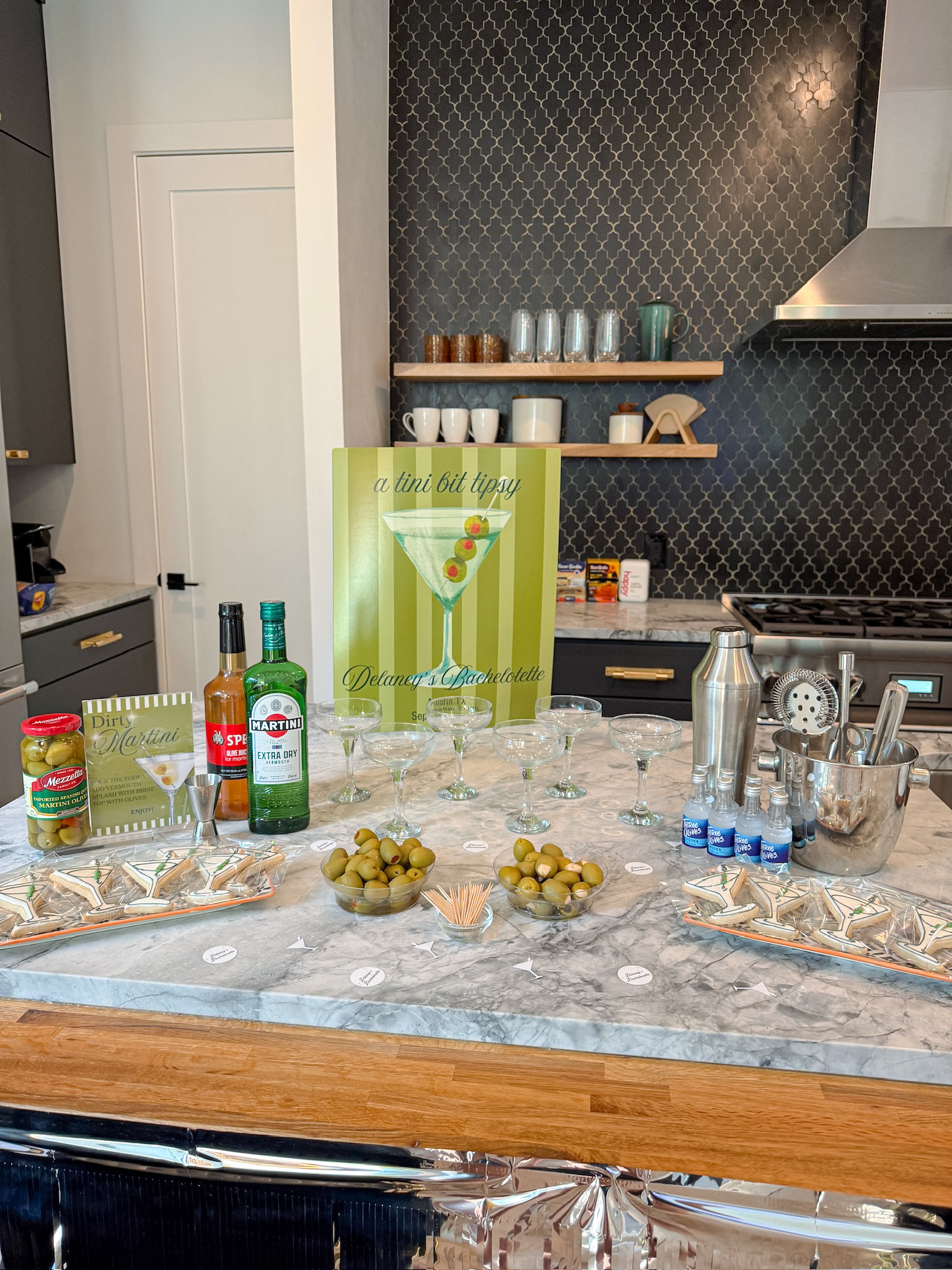 Martini-themed home bar on a marble kitchen island with coupe glasses, cocktail shaker, martini bottles and bowls of green olives set up for a bachelorette party in a modern kitchen.