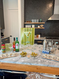 Martini-themed home bar on a marble kitchen island with coupe glasses, cocktail shaker, martini bottles and bowls of green olives set up for a bachelorette party in a modern kitchen.