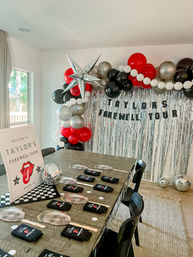 Indoor Austin farewell party setup with wooden table, disco balls, face paddle props and koozies, silver fringe backdrop, red-black-white balloon arch and metallic star balloon.