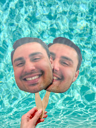 Hand holding two oversized smiling face cutout paddles on wooden sticks above turquoise swimming pool water — playful summer poolside prop