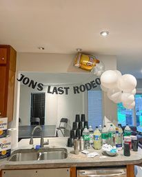 Home kitchen bar decorated for a party — black letter banner reading "LAST RODEO", a foil beer-can balloon and white balloon cluster above a counter lined with soda bottles, stacked plastic cups, Corona boxes and barware.