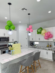 Festive modern white kitchen with marble island and gray bar stools, decorated with pink and green balloon clusters (including a lime-slice balloon), pendant lights, and a margarita-themed party bar setup.