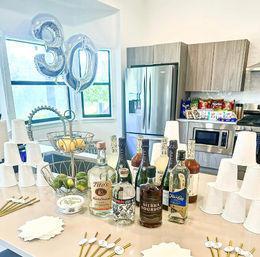 Modern home kitchen island set up for a 30th birthday: silver "30" balloons, bottles of spirits, stacked white cups, fruit tiers and snack bags.