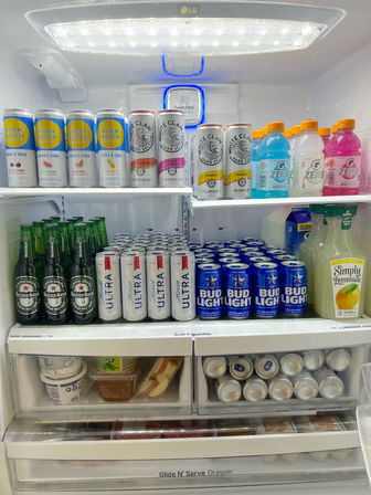 Open refrigerator neatly stocked with beverages: rows of canned vodka sodas and hard seltzers, colorful sports drink bottles and a lemonade jug, multiple beer cans and bottles, plus yogurt cups and sliced apples in the drawers.