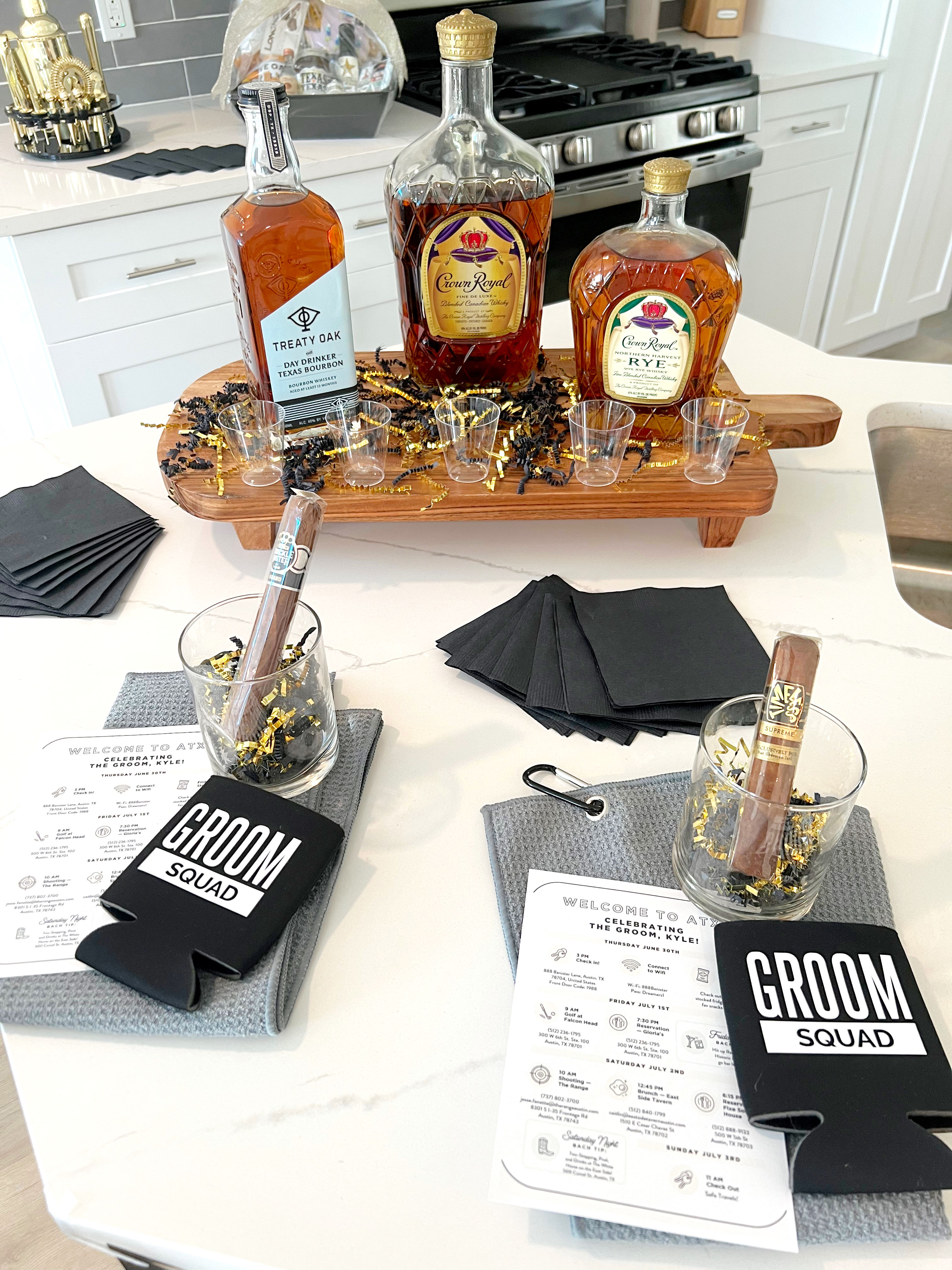 Groom squad welcome display on a white kitchen island: three whiskey bottles on a wooden serving board with shot glasses, cigars in glass tumblers with gold confetti, black napkins, 'Groom Squad' koozies and printed wedding weekend itineraries.