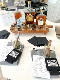 Groom squad welcome display on a white kitchen island: three whiskey bottles on a wooden serving board with shot glasses, cigars in glass tumblers with gold confetti, black napkins, 'Groom Squad' koozies and printed wedding weekend itineraries.