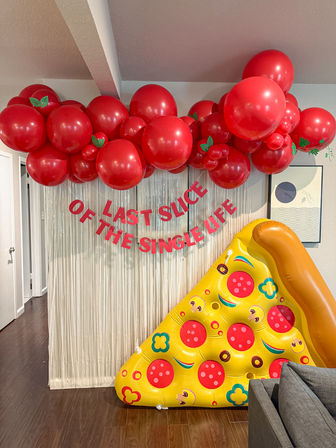 Indoor party backdrop: red balloon garland with green leaf accents, silver fringe curtain, pink banner reading 'LAST SLICE OF THE SINGLE LIFE', and oversized inflatable pizza slice on hardwood floor.