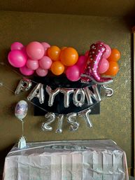 Indoor 21st birthday decorations: pink and orange balloon garland, silver foil letter balloons spelling a name with "21st", large pink foil number balloon and decorated table against gold patterned wallpaper.