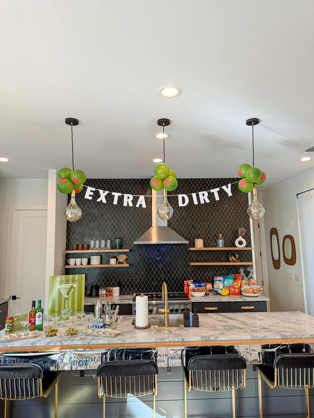 Modern home kitchen island with marble countertop and gold faucet, black tile backsplash, pendant lights with green balloon clusters and an "EXTRA DIRTY" banner, bar snacks and black velvet stools.