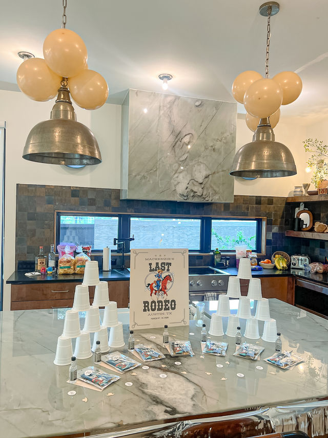 Marble kitchen island party setup with cowboy-themed “Last Rodeo” sign, stacked white plastic cups, mini liquor bottles, goody bags and clustered balloons beneath industrial pendant lights.