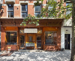 Wood-front boutique hotel entrance in a red-brick brownstone on a tree-lined city sidewalk with large display windows and hanging lights.