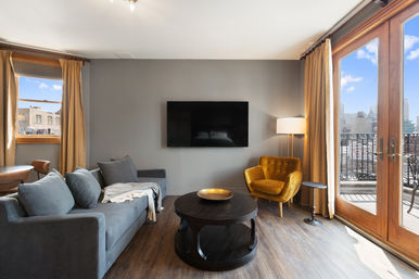 Cozy urban apartment living room with a gray sofa and throw, mustard velvet armchair, wall-mounted TV, round dark coffee table, floor lamp, and large French doors opening to a balcony with city skyline views.
