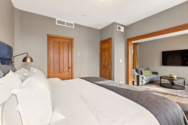 Bright modern bedroom in an urban apartment featuring a king bed with white linens and gray throw, brass reading lamp, wood doors and trim, neutral gray walls and hardwood floors opening to a cozy sitting area with sofa and wall-mounted TV.