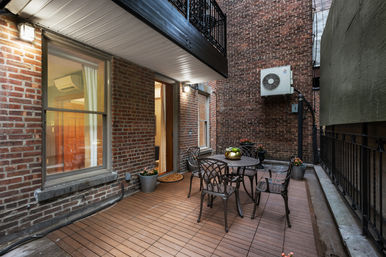 Cozy urban brick courtyard patio with metal dining table and chairs on wood-deck tiles, potted plants, wall-mounted AC unit and iron railing