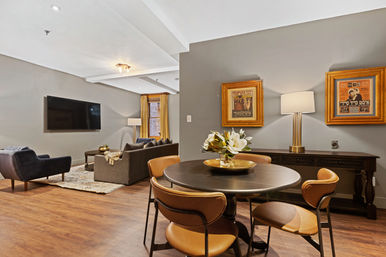 Cozy open-concept apartment living and dining area with hardwood floors — round black dining table with tan leather chairs and magnolia centerpiece, gray sofa and armchair facing a wall-mounted flat-screen TV, framed vintage posters and warm ambient lighting.