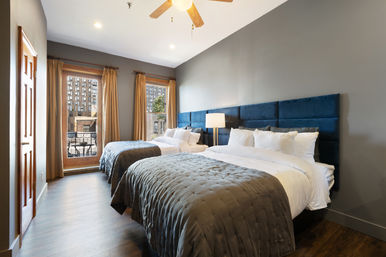 Bright urban bedroom with two queen beds, tufted navy-blue headboard, white linens and gray quilts, hardwood floors, ceiling fan, floor-to-ceiling windows with beige curtains opening to a small balcony and cityscape