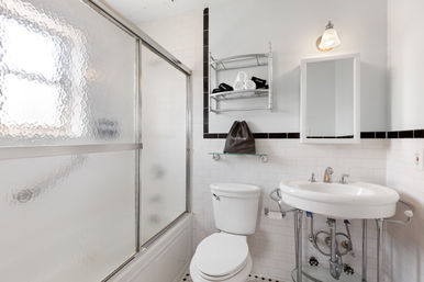 Bright white bathroom with frosted glass sliding shower door, white toilet and pedestal sink with exposed chrome plumbing, wall-mounted mirror cabinet, chrome shelving with rolled towels, and white subway tile with black trim.