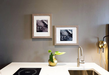 Stylish modern kitchen countertop with white marble island, stainless gooseneck faucet and undermount sink, small gold vase with green leaves, induction cooktop, brass wall sconce, and two framed black-and-white photos on a gray wall.