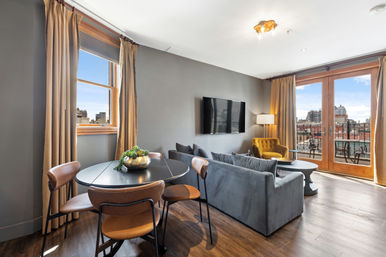 Modern urban apartment living room with gray sofa and mustard velvet armchair, wall-mounted TV, round black dining table with leather chairs, hardwood floors and French doors opening to a balcony with city skyline views