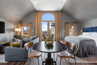 Airy urban loft suite with vaulted ceiling, king bed, cozy sofa seating, round dining table and arched balcony doors revealing a city skyline and rooftop water tower.