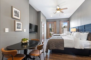 Cozy urban retreat studio with two plush beds and blue upholstered headboard, gray bedding, bedside lamp, wall-mounted TV, round dining table with leather chairs, ceiling fan, and balcony doors framing a downtown city skyline.