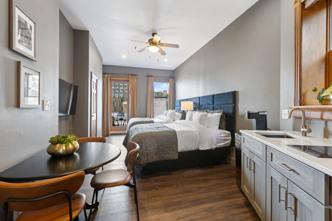 Modern downtown studio hotel room with two plush double beds and navy tufted headboard, gray bedding, kitchenette with sink, round dining table with leather chairs, ceiling fan, and balcony city view.