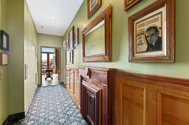 Sunlit urban hallway with green walls, patterned blue carpet, rich wooden wainscoting and vintage framed posters, leading through an open doorway to a bright dining area and city views.
