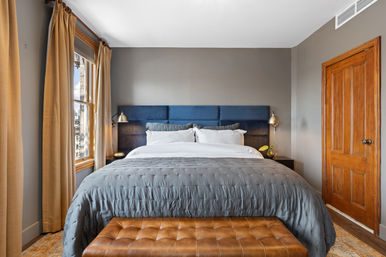 Cozy urban apartment bedroom with navy upholstered headboard, gray comforter, white pillows, tufted leather bench, wooden door and sunny window with beige curtains.