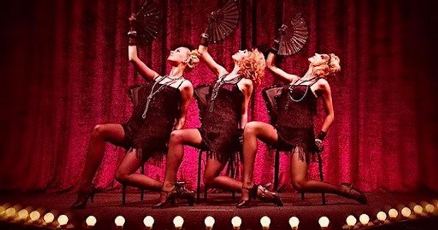 Three cabaret dancers in black flapper fringe dresses and stockings strike synchronized seated poses with black fans on a red-curtained theater stage lined with marquee bulbs.