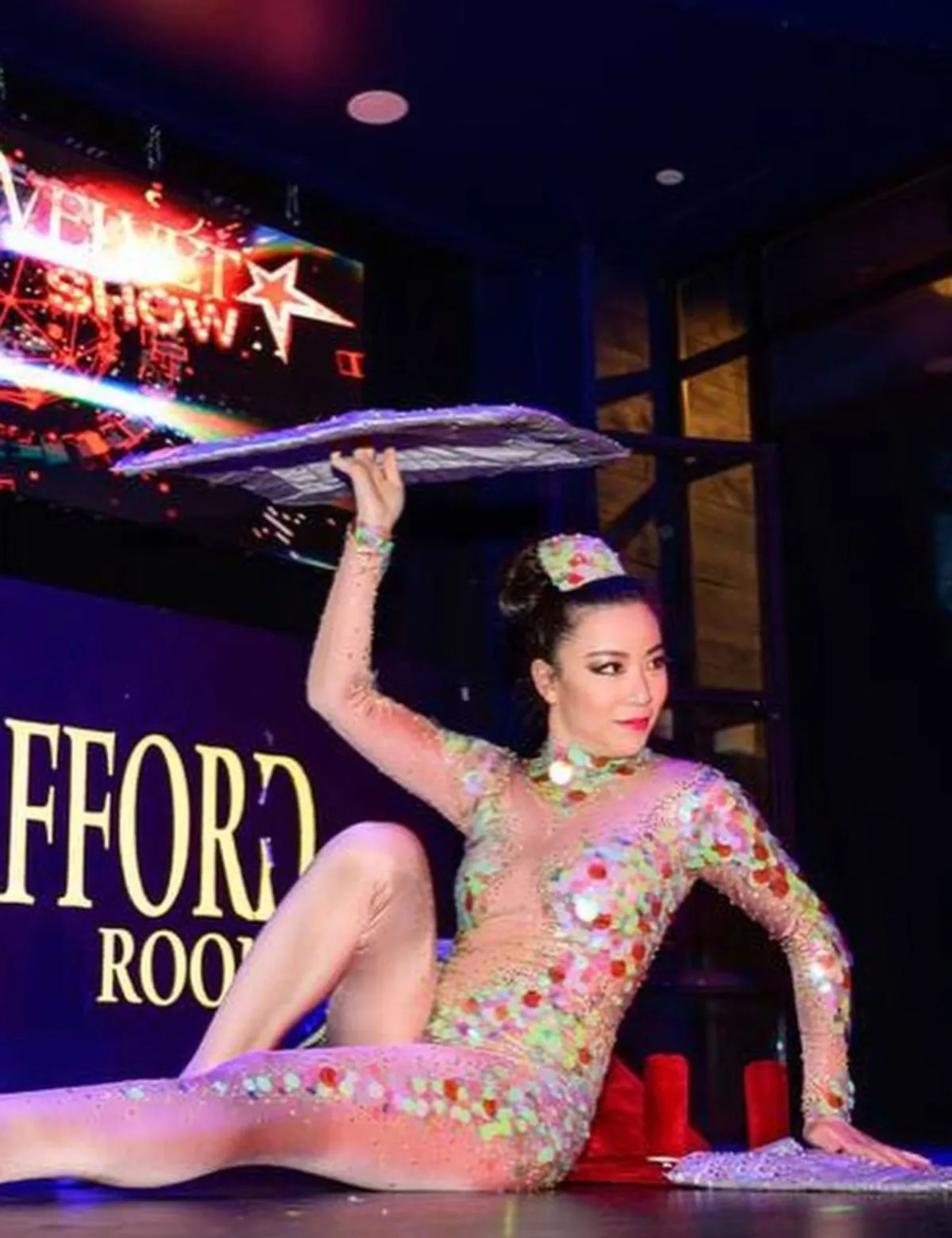 Cabaret performer in an iridescent sequin bodysuit and matching headpiece balances a mirrored tray above her head while posing on a brightly lit stage.
