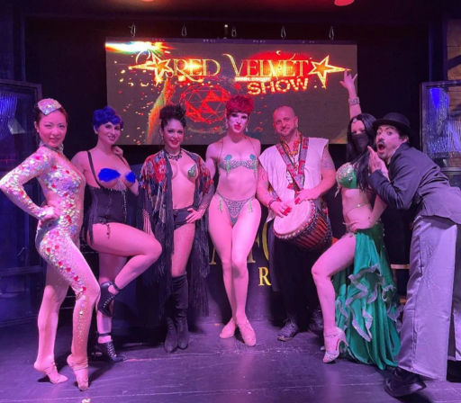 Seven burlesque and variety performers in colorful costumes — sequins, pasties, fringe, bikini-style outfits and a belly-dance skirt — posing with a drummer on a cabaret stage under a glowing marquee.