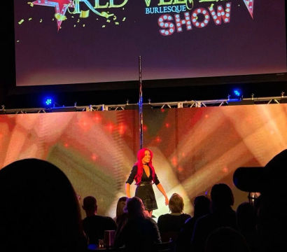 Live burlesque performer with bright red hair on a lit theater stage next to a vertical pole, colorful marquee-style projection and star graphics above, audience silhouettes in the foreground.