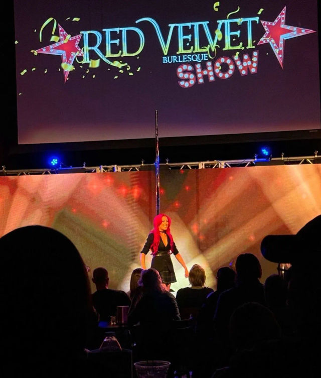 Live burlesque performer with bright red hair on a lit theater stage next to a vertical pole, colorful marquee-style projection and star graphics above, audience silhouettes in the foreground.
