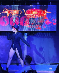 Burlesque performer in a top hat and tailcoat strikes a dramatic pose on a nightclub cabaret stage under a glowing marquee, purple-blue lighting and seated audience silhouettes in the foreground.
