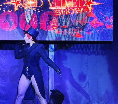 Burlesque performer in a top hat and tailcoat strikes a dramatic pose on a nightclub cabaret stage under a glowing marquee, purple-blue lighting and seated audience silhouettes in the foreground.