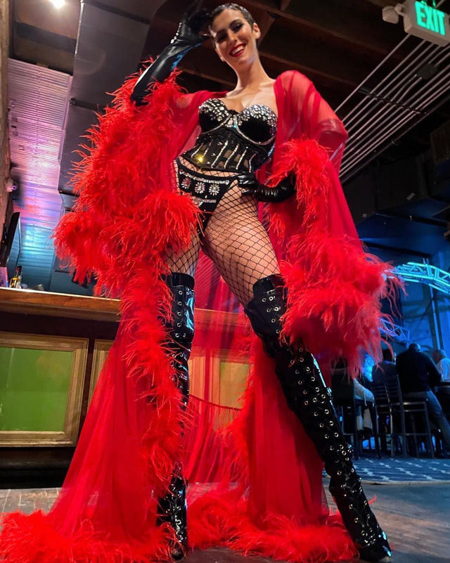 Burlesque performer in a red feathered robe and bejeweled black corset, fishnet stockings and thigh-high patent lace-up boots, striking a confident pose on a nightclub stage near the bar.