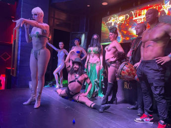 Burlesque and cabaret performers on a nightclub stage during a live show — singer with short platinum hair at left, costumed dancers in sequins, feathers and fringe, a male performer doing the splits, colorful LED backdrop and stage lights.