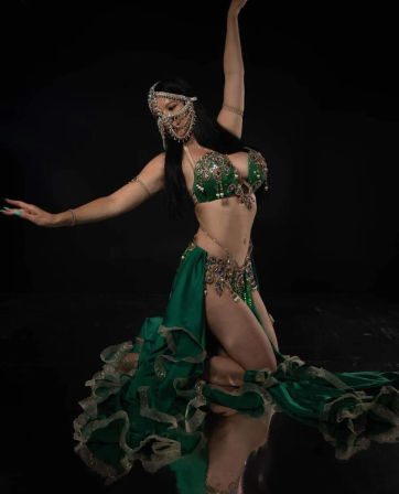 Belly dancer in a green jeweled costume and beaded face veil kneeling on a reflective black studio floor, striking an elegant arm-up pose with flowing ruffled skirt.