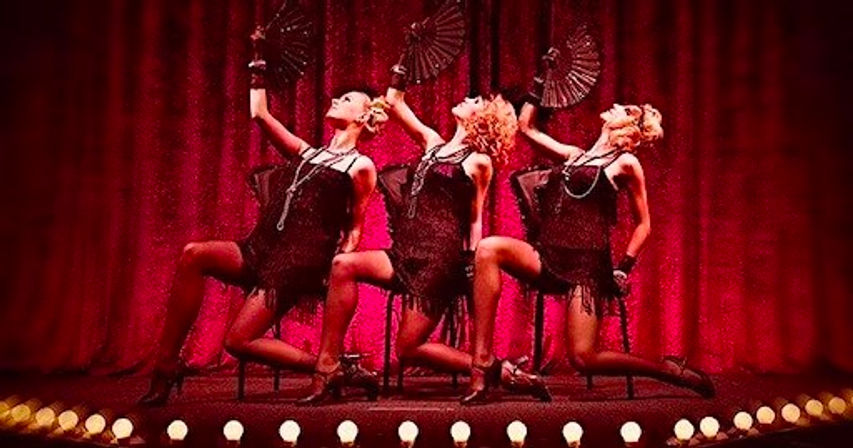 Three cabaret dancers in black fringe dresses and pearls strike synchronized poses with handheld fans on a theater stage before red velvet curtains and glowing footlights.